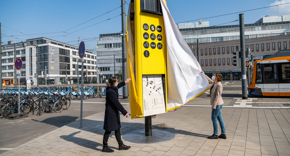 Zwei Frauen stehen auf einem Bahnhofsvorplatz und enthüllen eine große gelbe Informationssäule