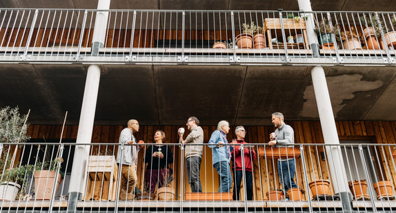 Sechs Personen unterhalten sich auf einem Balkon eines mehrstöckigen Gebäudes mit sichtbaren Betonstützen und Pflanzgefäßen