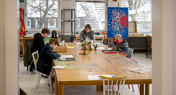 An einem Tisch großen Tisch in einer Kunstwerkstatt arbeiten Künstler an Bildern.