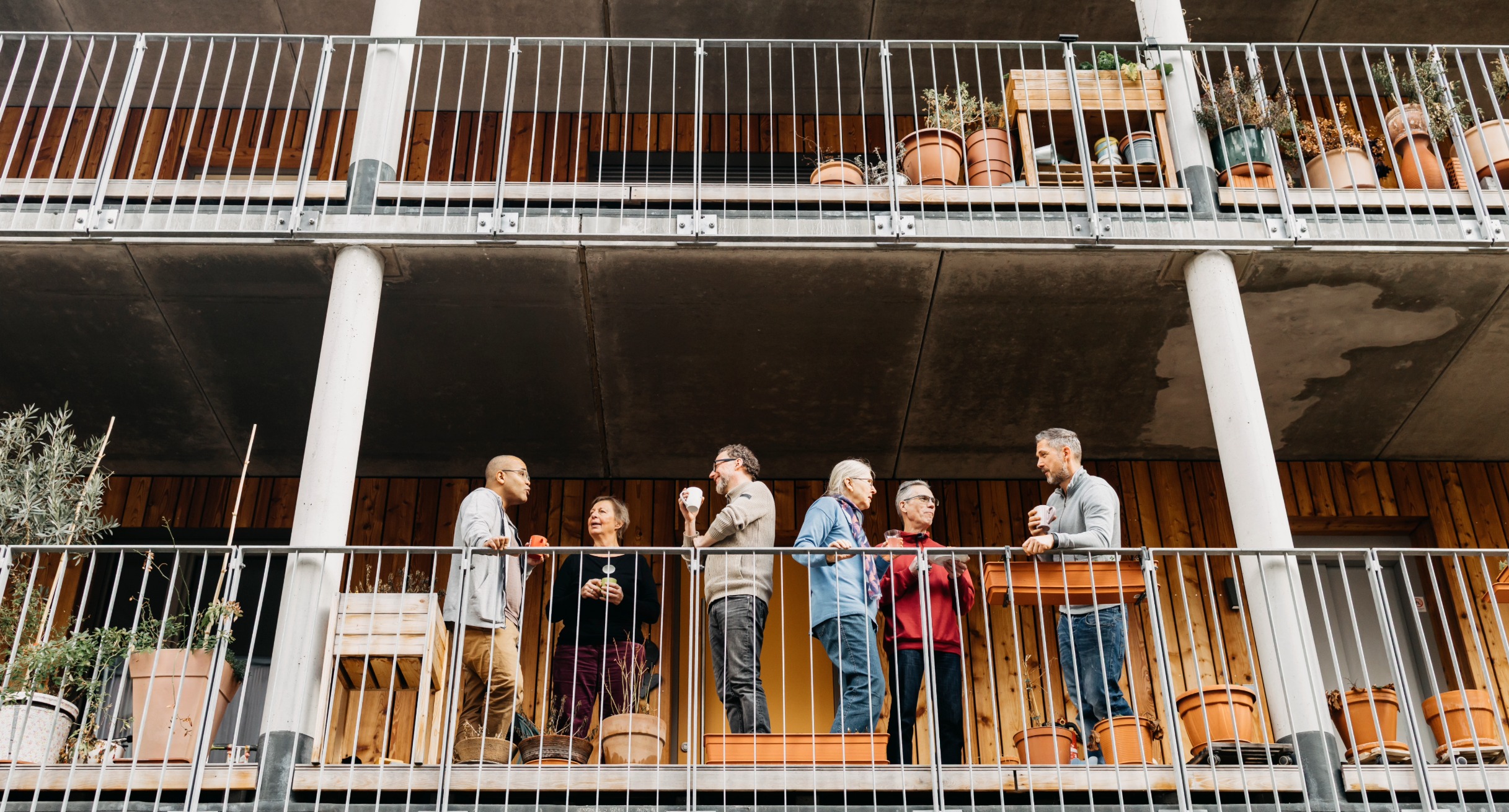 Sechs Personen unterhalten sich auf einem Balkon eines mehrst&ouml;ckigen Geb&auml;udes mit sichtbaren Betonst&uuml;tzen und Pflanzgef&auml;&szlig;en