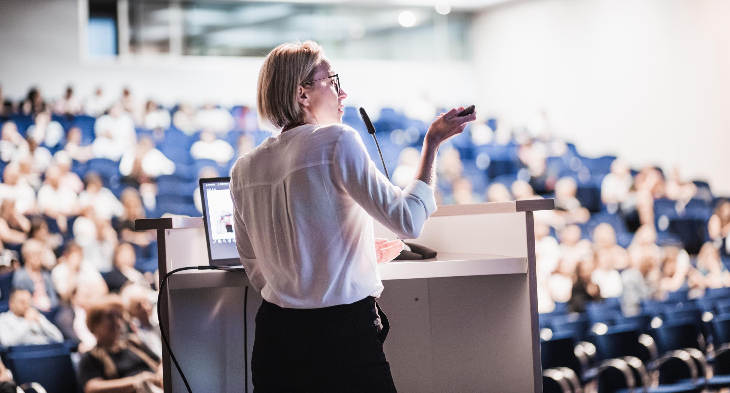 Female speaker giving a talk on corporate business conference. Unrecognizable people in audience at conference hall. Business and Entrepreneurship event.']