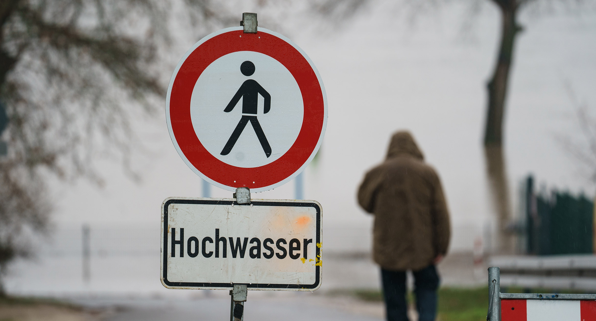 Ein Mann geht hinter einem Schild mit der Warnung „Hochwasser“ zu einem Flussufer.']