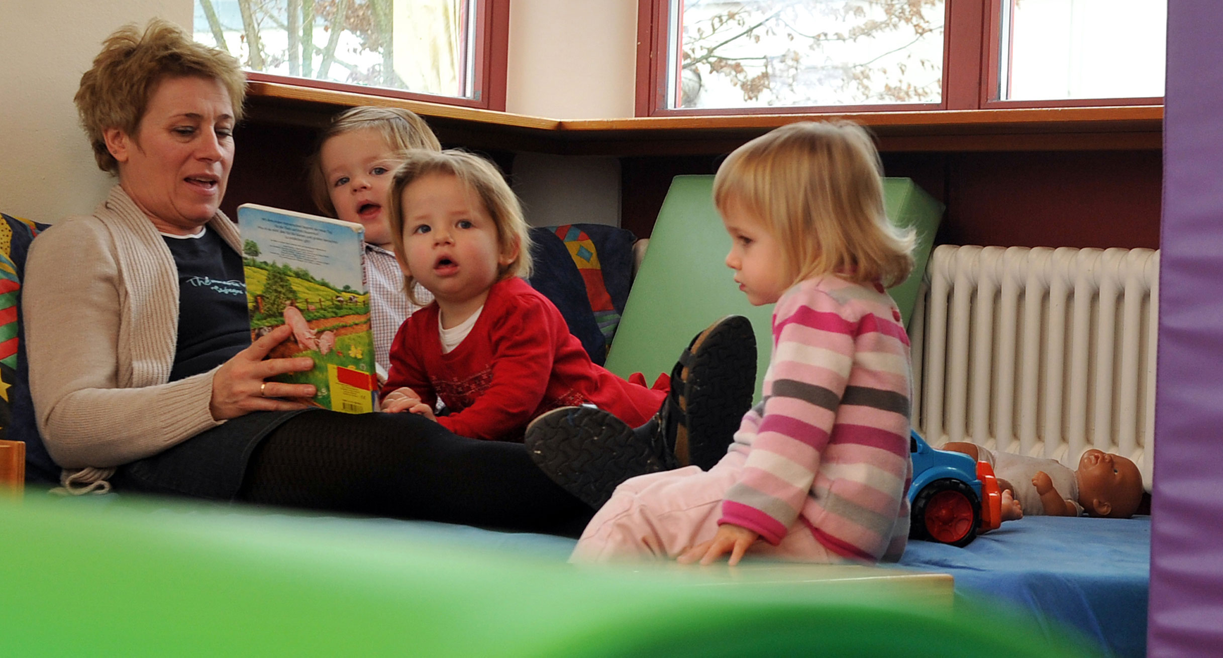 Eine Erzieherin ließt drei Kleinkindern aus einem Buch vor (Bild: © dpa).']