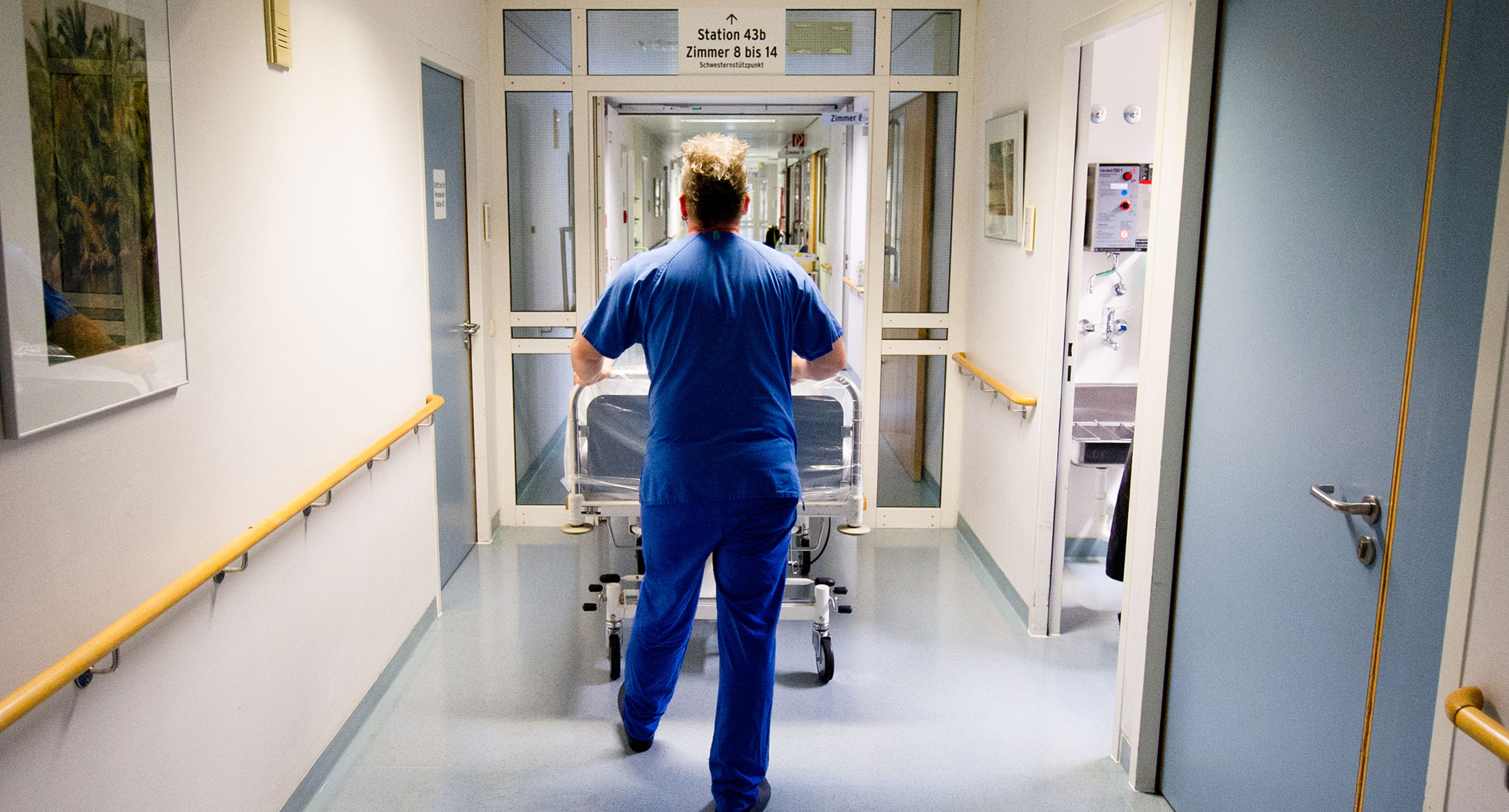 Ein Krankenpfleger schiebt in einer Klinik ein Krankenbett über einen Flur. (Foto: © dpa)']