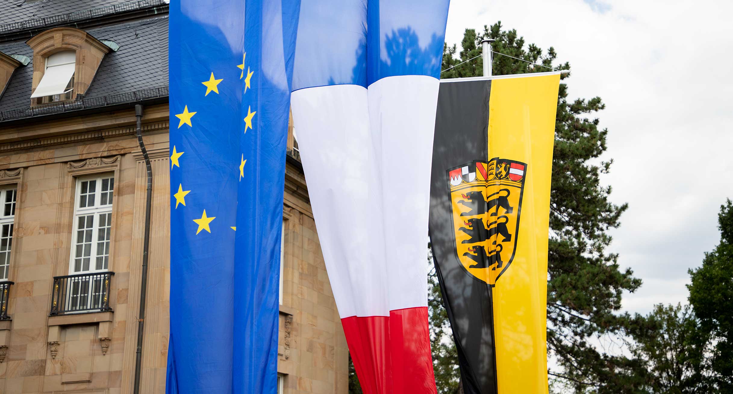 An Fahnemasten hängen die Fahne der EU, die französische Fahne und die baden-württembergische Fahne.']