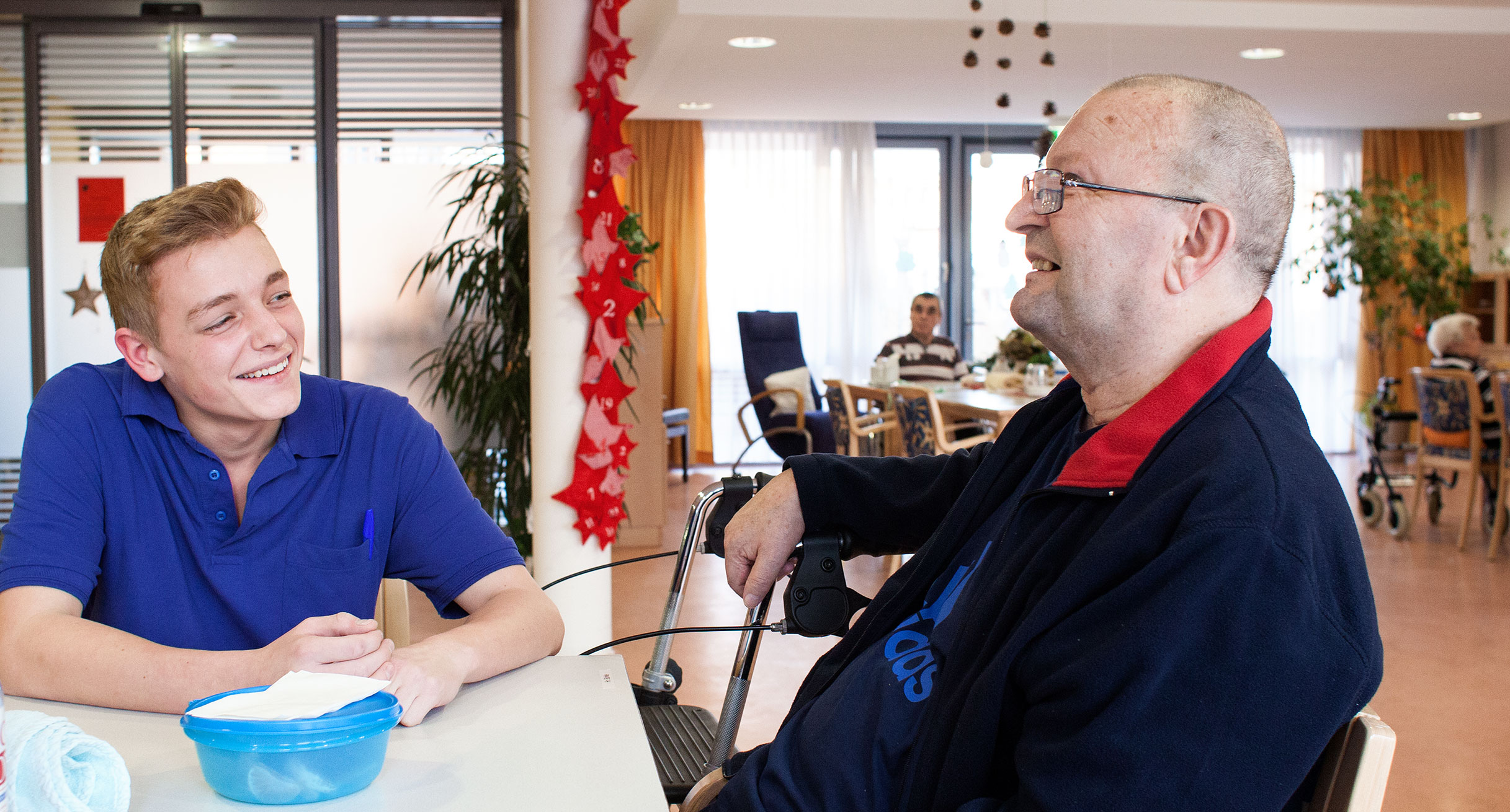 An einem Tisch im Pflegeheim ist ein junger Pfleger im Gespräch mit einem älteren Mann.