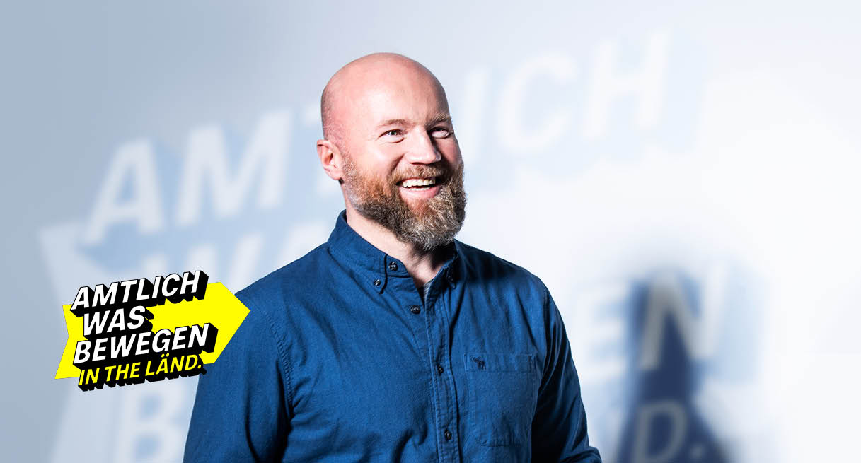 Lächelnder Mann in blauem Hemd vor dem Slogan "Amtlich was bewegen in THE LÄND."