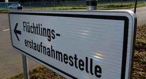 Ein Schild mit der Aufschrift &bdquo;Fl&uuml;chtlingserstaufnahmestelle&ldquo;, im Hintergrund ein Fahrzeug an einer Einfahrt.