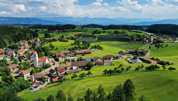 Das beschauliche Dorf Hiltensweiler, ein Teilort von Tettnang, wird von der Abendsonne angestrahlt. Im Hintergrund sind der Bodensee und die Alpen zu sehen.