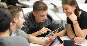 Drei jugendliche Berufssch&uuml;ler und eine l&auml;chelnde Berufssch&uuml;lerin erkunden gemeinsam die Funktionsweise eines Tablets, das einer der Sch&uuml;ler in der Hand h&auml;lt.
