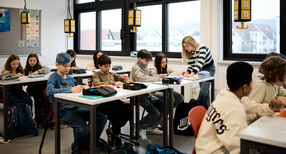 Blick in ein Klassenzimmer, in dem Sch&uuml;lerinnen und Sch&uuml;ler an Tischen sitzen. Eine Lehrerin steht neben einem der Tische.