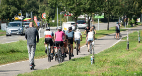 Zahlreiche Menschen sind auf einem Radweg bei Sonnenschein auf ihren Fahrr&auml;dern und E-Roller unterwegs.