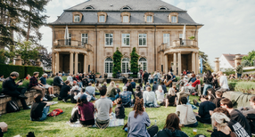 Menschen sitzen auf dem Rasen vor der Villa Reitzenstein und h&ouml;ren einer Person zu.