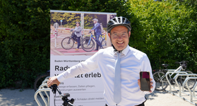 Staatssekret&auml;r Raimund Haser steht vor seinem Fahrrad mit einem Fahrradhelm auf dem Kopf und h&auml;lt ein Handy in die Kamera.