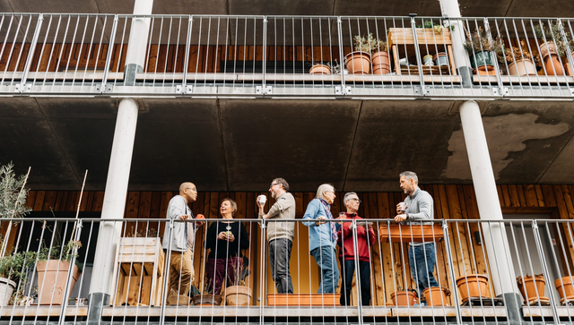 Sechs Personen unterhalten sich auf einem Balkon eines mehrst&ouml;ckigen Geb&auml;udes mit sichtbaren Betonst&uuml;tzen und Pflanzgef&auml;&szlig;en