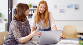 In einem B&uuml;ro unterhalten sich zwei Frauen. Eine sitzt an einem Tisch und eine steht neben dem Tisch. Auf dem Tisch steht ein Laptop.
