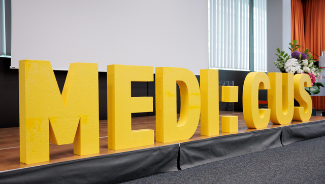 Schriftzug &bdquo;MEDI:CUS&ldquo; auf einer B&uuml;hne