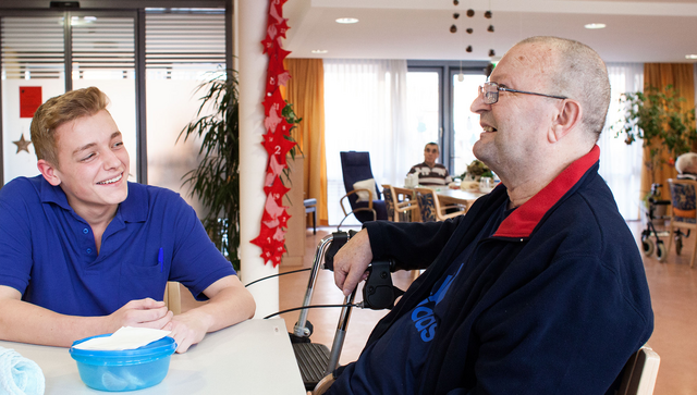An einem Tisch im Pflegeheim ist ein junger Pfleger im Gespr&auml;ch mit einem &auml;lteren Mann.