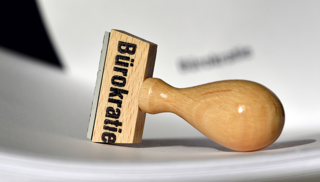 Ein Stempel mit der Aufschrift "B&uuml;rokratie" liegt auf Papieren.