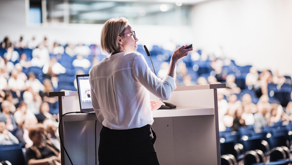Female speaker giving a talk on corporate business conference. Unrecognizable people in audience at conference hall. Business and Entrepreneurship event.